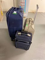 Three pieces of wheeled luggage including two navy blue and one beige suitcase standing on a concrete floor next to a water heater.