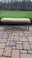 Metal patio bench with cushion on a stone patio, showing side view of the bench and cushion.