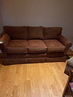 Front view of brown three-seater fabric sofa showing three seat cushions and three back cushions, rounded armrests, and skirted base.