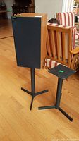 Speaker with wood casing and black fabric grille standing on one metal tripod base speaker stand, two metal stands shown, hardwood floor background