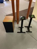 Photo of one wooden finish speaker with black fabric front and two black metal speaker stands on basement floor