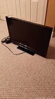 Front view of the 32-inch Insignia TV showing the black screen, frame, and attached power cord lying beside it on carpeted floor.