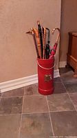 Umbrella stand with multiple canes and umbrellas visible inside, positioned on tiled floor against beige wall.