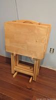 Four solid wood TV trays standing upright in the storage stand. The trays have a natural wood finish with a cutout handle at the top.