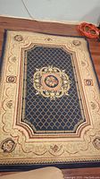 Full view of decorative area rug on wooden floor showing dark blue center with lattice and floral medallion, cream border with flowers.