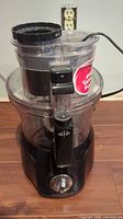 Front view of the black food processor showing base, clear bowl, control knob, and power cord.