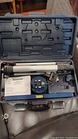 Open black plastic carrying case containing a silver adjustable tripod, blue Mastercraft laser level unit, and instruction manual.