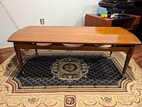 Full front view of the mid century modern solid wood coffee table showing the curved top, tapered legs, and decorative apron, sitting on a patterned rug.