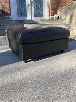 Side view of black leather ottoman showing cushioned top and base with legs.
