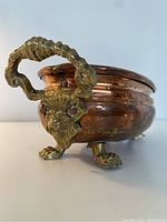 Close side view showing lion head brass handle and claw foot on copper jardiniere planter with some wear and dents on patina.