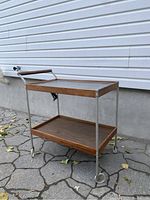 Full left angle view of the serving cart showing teak veneer shelf, metal frame, glass top and castor wheels.