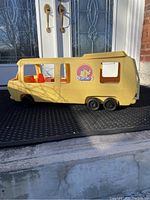 Side view showing yellow plastic camper exterior with large Barbie logo sticker and three visible wheels with black tires and red interior seats.