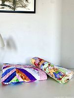 Two folded quilts displayed side by side. The quilt on the left has a colorful diagonal stripe pattern with shiny fabrics, the one on the right is rolled and shows nursery rhyme themed prints.