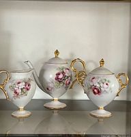 Full view of the three-piece porcelain tea set with painted roses and gold trim, showing front and side of teapot, creamer, and sugar bowl.