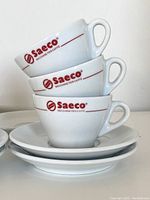 Stacked close-up view of three white ceramic Saeco cappuccino cups with red logo and text 'Macchine Per Caffe' on each cup, showing handles and rims, alongside three matching saucers underneath.