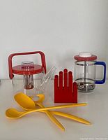 Front view of the Bodum tea pot, Pyrex French press, red hand-shaped holder, and three yellow melamine utensils on a white surface.