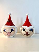 Pair of round vintage ceramic candy jars shaped as Santa Claus and Mrs. Claus heads, each with removable tall red Santa hat lids, on white surface