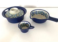 Three pieces of ceramic cookware including casserole with lid, saucepan and small sauce pot with long handles, showing floral painted interiors and matte blue exteriors.