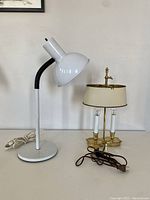Both lamps shown side-by-side on a table. White gooseneck table lamp on the left with power cord coiled, brass bouillotte lamp on the right with brown power cord and plug.