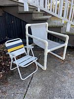 Two chairs on outdoor concrete surface; white child's folding chair with colored slats and white PVC pipe armchair with vinyl sling seat and back, placed side by side.