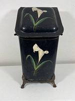 Front view of black tole painted metal coal scuttle with white lily design and cast iron feet on a white background.