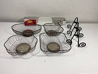 Four wire bread baskets with metal wire construction and round bases, three black metal wall-mounted plate racks with curled base design, unopened 3M Command stick-on wall shelf in packaging on a white surface against plain background.