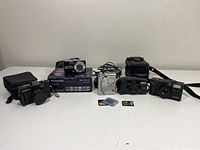 Lot containing five cameras placed on a white surface, including the Panasonic Lumix DMC TZ3 with box, Yashica Elite 70 Zoom, Vivitar DL50, FujiFilm FinePix 4700, and Ultronic camera with accessories and batteries.