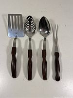 Front view of four vintage Cutco serving utensils laid flat showing wooden handles and stainless steel heads: slotted spatula, perforated spoon, solid spoon, and fork.