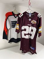 Two jerseys hanging side by side, left white/black/orange Flyers hockey jersey with number 22, right burgundy Redskins football jersey number 29 with 70th anniversary patch.