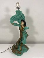 Full front view of vintage chalkware lamp with female figure in teal and gold outfit standing on rocky base, flowing fabric overhead, lamp socket at top.