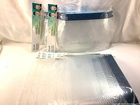 Photo showing several face shields sealed in clear plastic packaging with white tabs containing product info, stacked together.