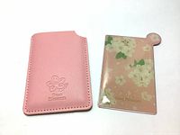 Pink floral hand mirror next to its matching pink faux leather protective case with embossed logo.