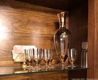 Full display of the vintage decanter with stopper, six matching glasses, and gold-colored napkin holder with napkins on a glass shelf.