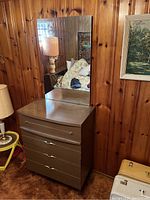 Front view of wooden dresser with attached rectangular mirror in a wood-paneled room with a lamp and paintings nearby.