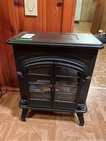 Front view of the black metal electric fireplace showing double glass doors with grid pattern, top lid, and legs.