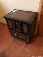 Single wood veneered particle board end table with three drawers and decorative metal handles, placed on wood floor near wall