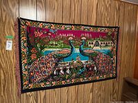 Full view of the fabric wall hanging showing the peacock design and floral border hung on a wooden-paneled wall.