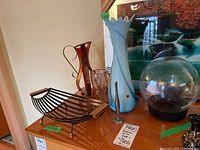 View of the full lot including the tall blue Italian art glass vase with applied threading, red and amber glass vase, clear faceted vase, metal bud vase, clear globe, black metal rack, and small basket on a wooden surface