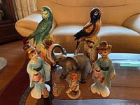 Eight Japanese ceramic figurines including elephant, two birds, two women in traditional attire, small child, and two Wade figurines arranged on a living room table and couch.