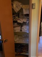 Open linen closet shelves with stacks of folded embroidered linens, doilies, and vintage fabrics in cream, white, and pastel colors