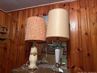 Two Mid-Century Modern lamps with fabric shades shown against wood panel wall and mirror, one with white glass base and beige fabric shade, one with beige ceramic composite base and orange woven fabric shade.