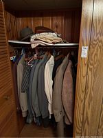Closet view showing multiple men's suits and jackets hung, shirts stacked on shelf, brown leather belt, and brown hat on shelf