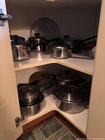 Corner cabinet interior showing multiple stainless steel pots and pans with lids, some stacked, mostly with black handles and knobs.