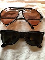 Both pairs of sunglasses laid flat on fabric background, one black framed Ray-Ban Wayfarer, one metal frame Serengeti Drivers with brown lenses.