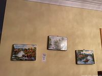 Three rectangular paintings with landscape and river scenes displayed on a wall