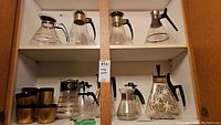 Full cabinet shelf image showing eight vintage glass coffee carafes with black handles and golden accents, some having painted floral motifs and some paired with gold and black coffee cups.