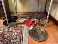 Overview of 11 trays on floor beneath a metal-framed table, showing various sizes, shapes and colors including a bright red floral tray and several silver plated trays.