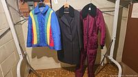 Full view of three outerwear items hanging: rainbow jacket, black trench coat, and burgundy ski suit.
