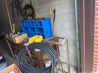 Overview of garage corner showing blue metal storage organizer, coiled black hose, vise on wooden bench, and garden tools leaning against wall.
