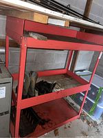 Red metal workshop storage rack with three shelves, Kamik boots and work shoes on middle shelf, wooden planks on upper shelf, garage background.